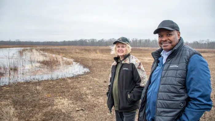 Two people standing by a wetland area in an open field on a cloudy day